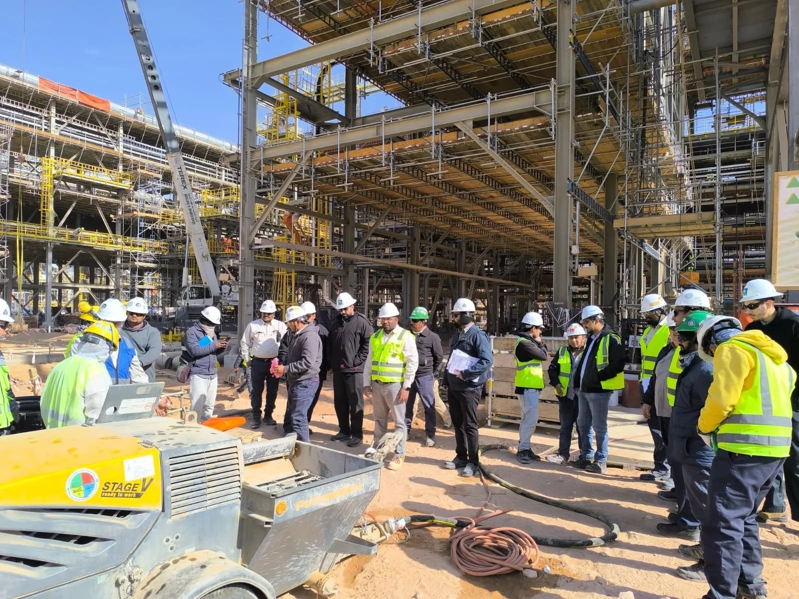 Industrial workers in helmets and high-visibility vests gathered beneath scaffolding at a project site.